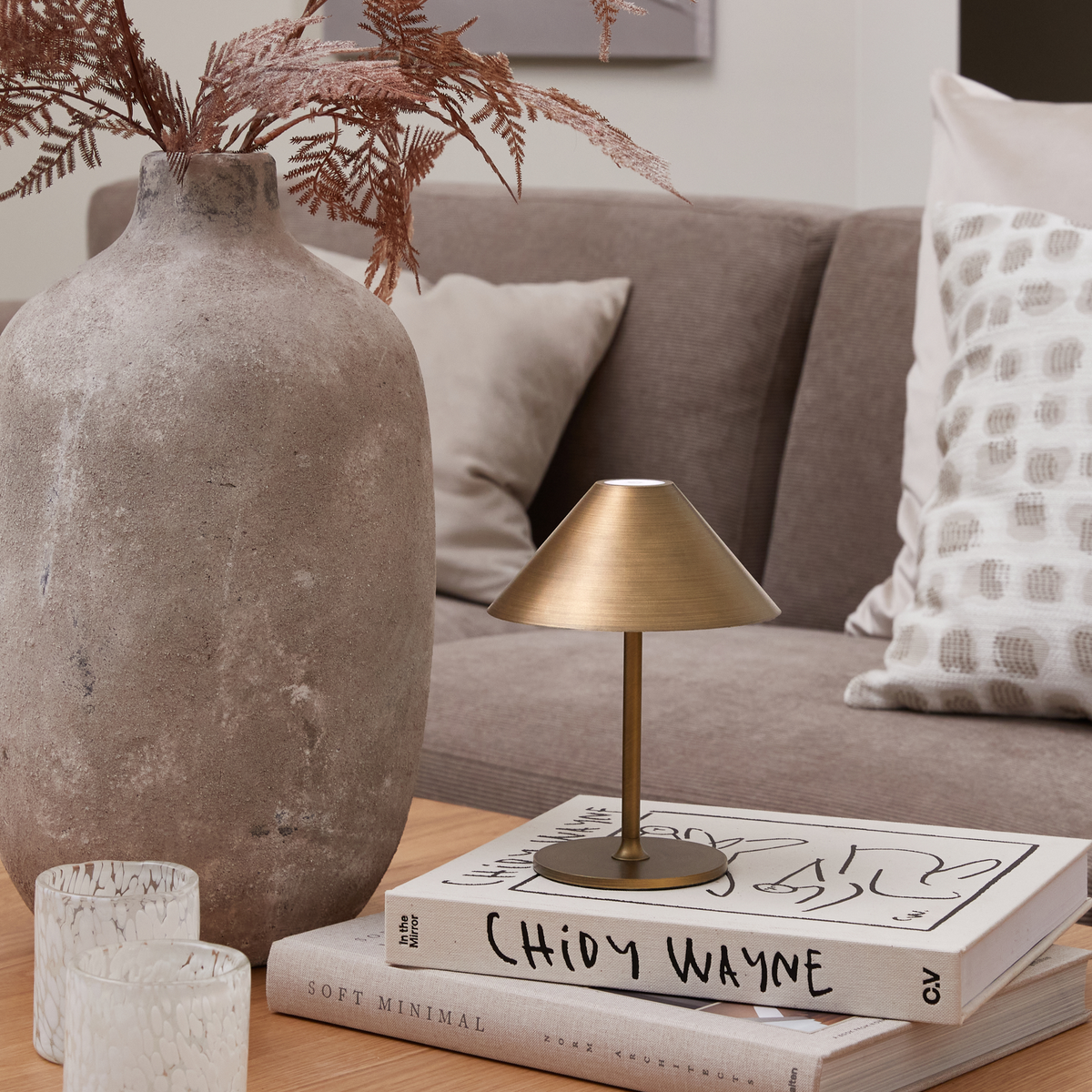 Nærbillede af en neutral stue: en lille messinglampe står på bøgerne ”Chidy Wayne” og ”Soft Minimal” ved siden af en stor stenvase med tørrede grene og to glasfyrfadsstager på et træbord, med beige sofa og puder i baggrunden.