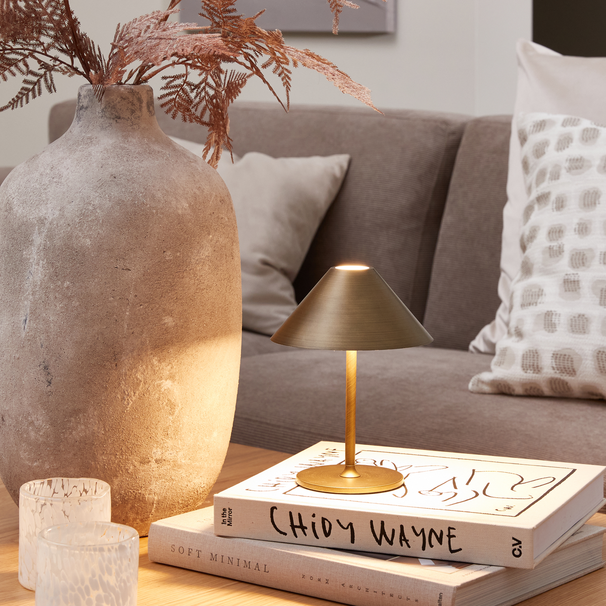 Lille messingfarvet bordlampe på to bøger, Chidy Wayne og Soft Minimal, ved siden af en stor rustik vase, fyrfadslys og en beige sofa med puder.