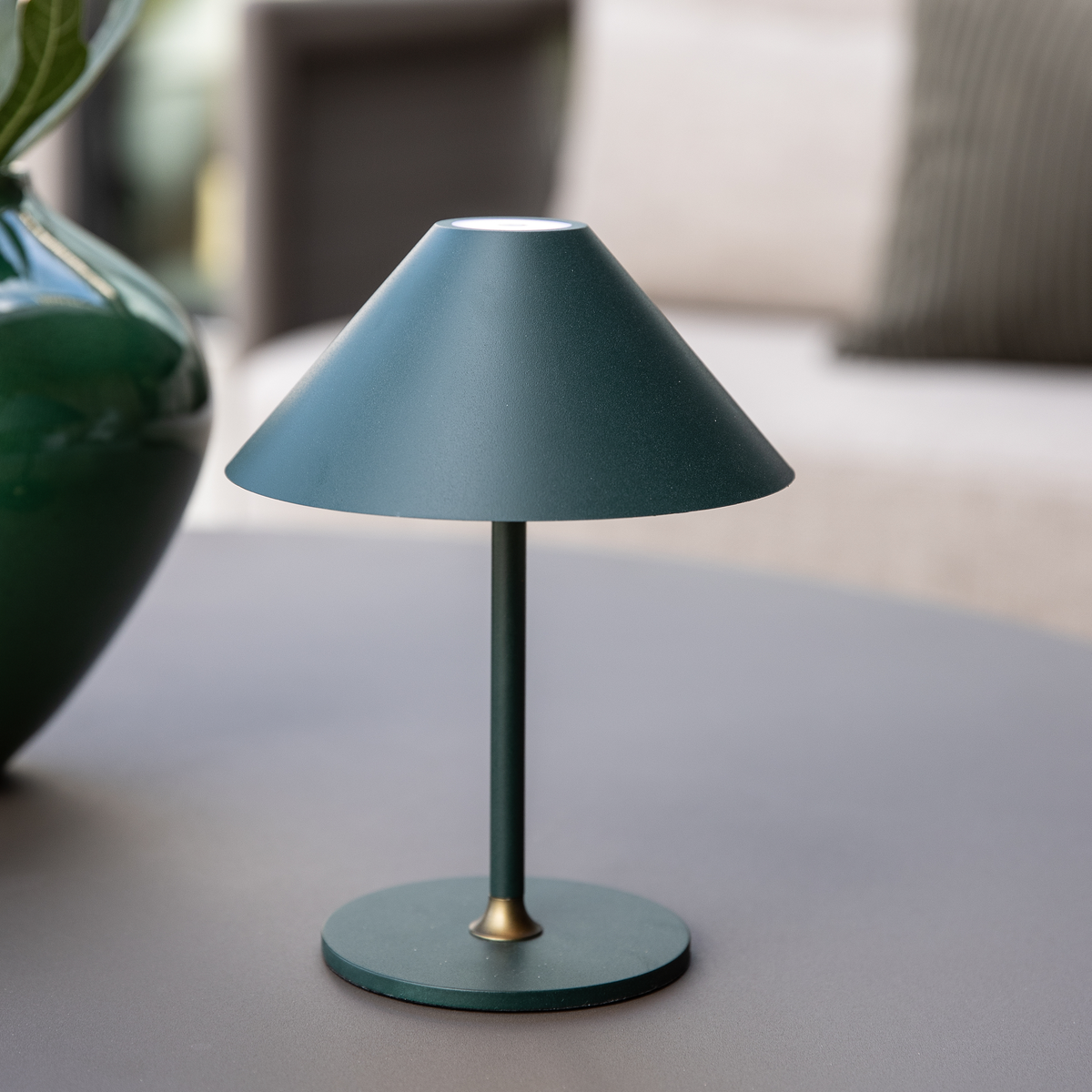 Grøn bordlampe med konisk skærm og rund fod på et sofabord, med en sløret vase i baggrunden.