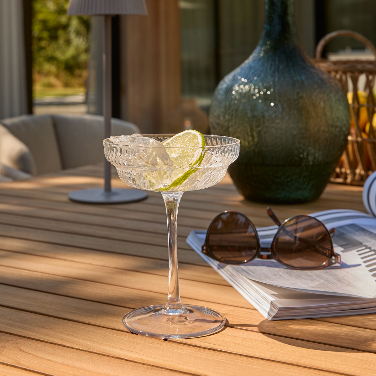 Cocktailglas i krystal med is og limeskive på udendørs træbord ved siden af solbriller, magasin og grøn glasvase i solskin.