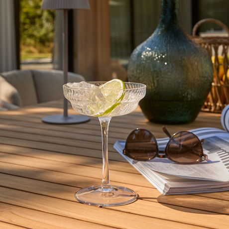 Cocktailglas i krystal med is og limeskive på udendørs træbord ved siden af solbriller, magasin og grøn glasvase i solskin.