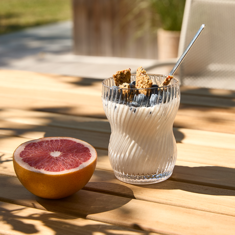 Glas med yoghurt toppet med blåbær og granola med ske ved siden af en halveret grapefrugt på et solbeskinnet udendørs træbord.