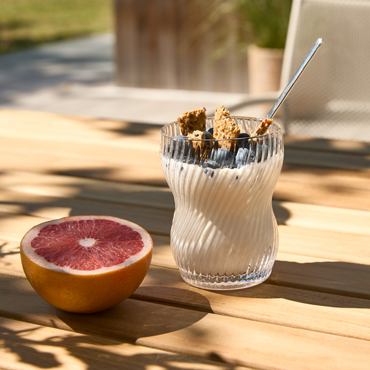 Glas med yoghurt toppet med blåbær og granola med ske ved siden af en halveret grapefrugt på et solbeskinnet udendørs træbord.