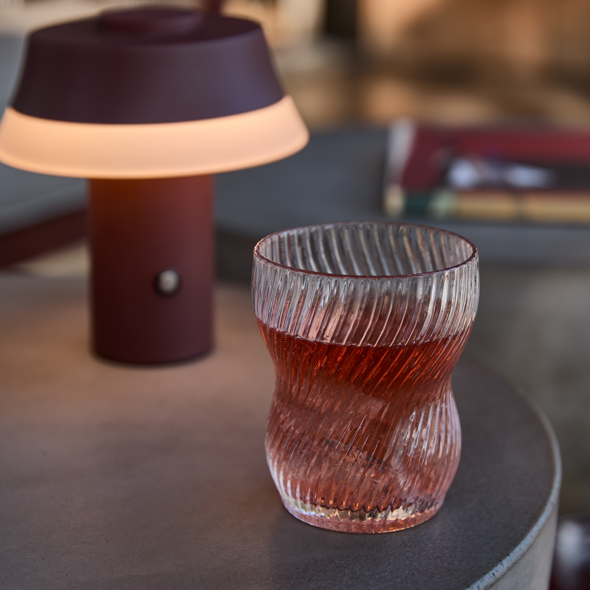 Riflet drikkeglas med rød drik på et rundt bord ved siden af en lille vinrød, bærbar lampe med varmt lys.