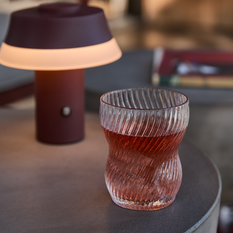Riflet drikkeglas med rød drik på et rundt bord ved siden af en lille vinrød, bærbar lampe med varmt lys.