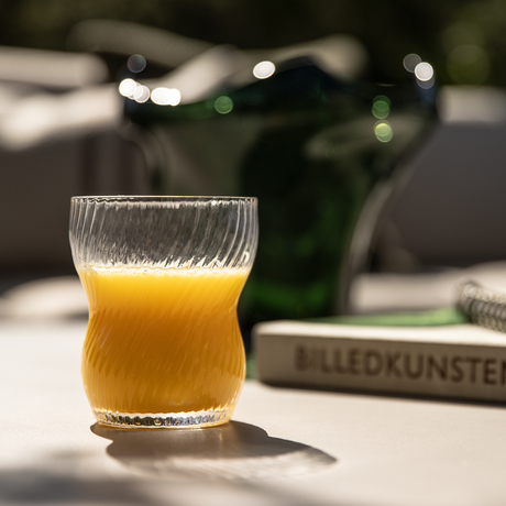 Glas med appelsinjuice på solrig terrasse, bølget riflet glas, sløret grøn kande i baggrunden og en bog, BILLEDKUNSTEN.