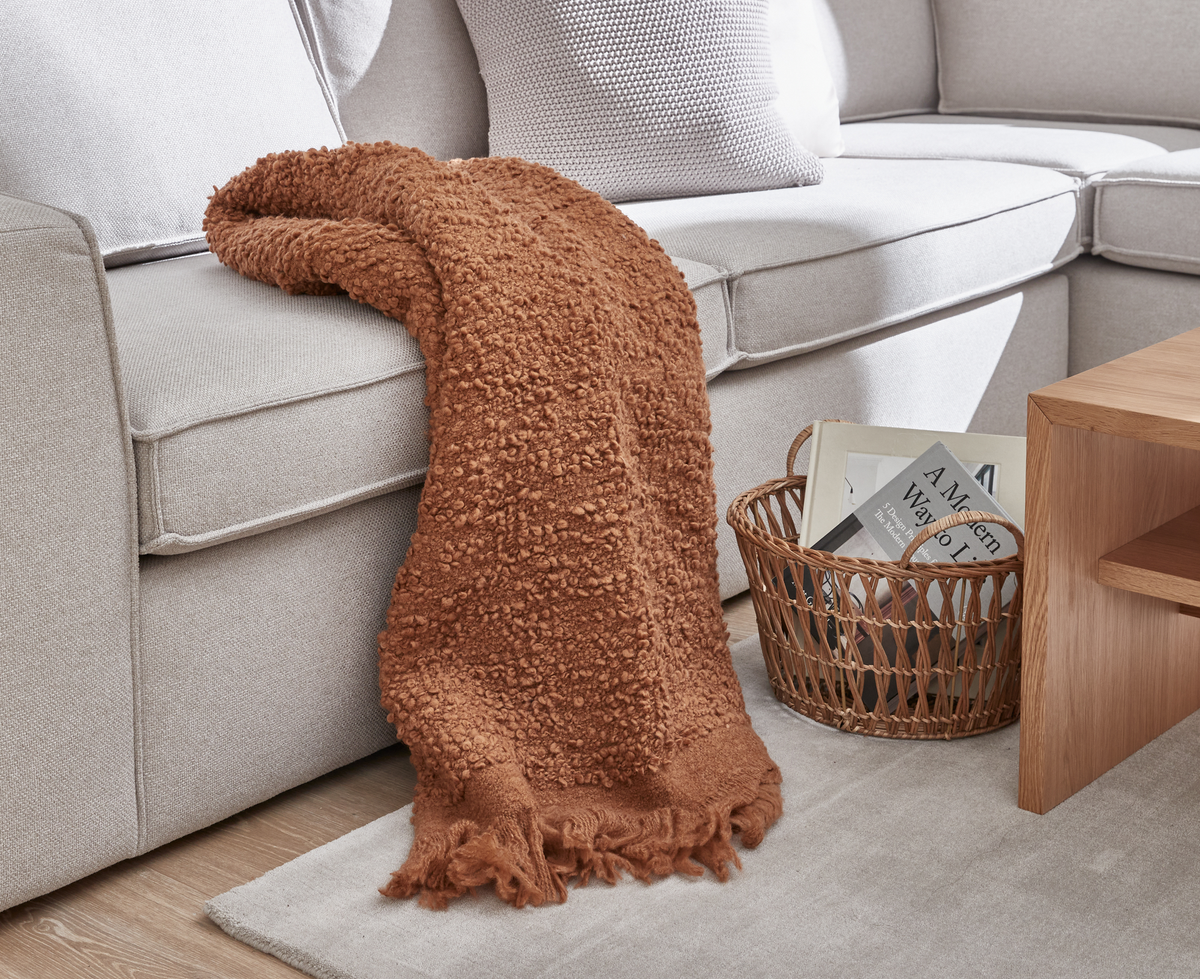 Beige sofa med rustfarvet, tekstureret plaid, der falder ned på et lyst tæppe; ved siden af står en fletkurv med en bog med titlen ”A Modern Way to...” og et sidebord i lyst træ.