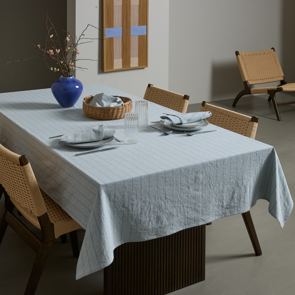 Minimalistisk spisescene med lyseblå stribet dug på et rektangulært bord dækket med tallerkener, stofservietter og riflede glas, fletstole omkring, blå vase med grene og fletkurv på bordet samt moderne vægkunst og loungestol i baggrunden.