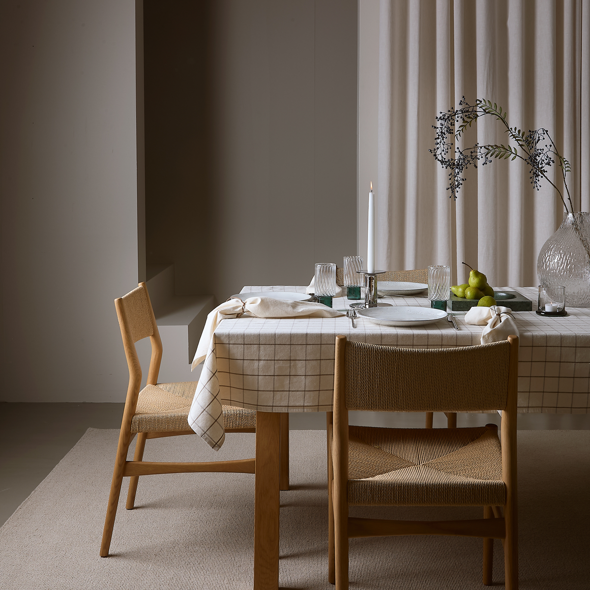 Stilrent spiseområde med to træstole, ternet dug, opdækning med glas og tallerkener, et slankt stearinlys, glasvase med gren og grønne pærer på bakke foran lyse gardiner.