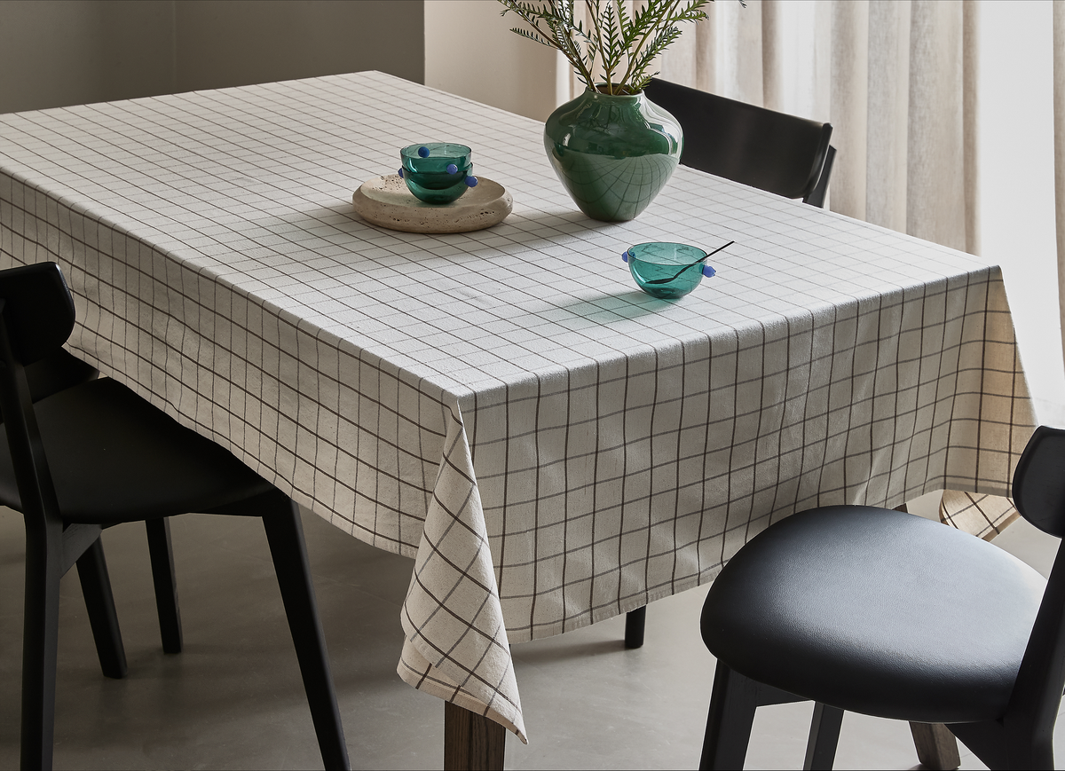 Spisebord med beige ternet dug, sorte stole, grøn vase med grene og to turkise glasskåle i blødt dagslys.