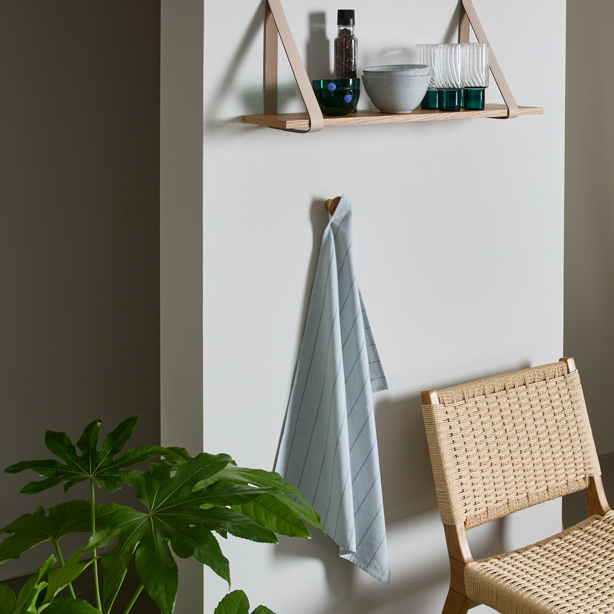 Væg med lille træhylde med læderremme, glas og skål samt en flaske; lyst stribet viskestykke hænger på knage ved siden af en flettet stol og en grøn stueplante.
