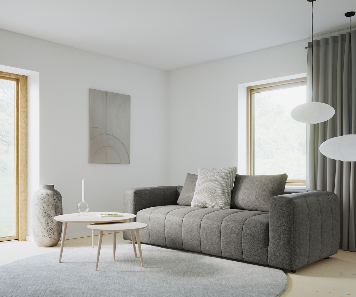 Lyst stue med grå sofa, to lyse sofaborde, rundt gråt tæppe, høje gardiner og rispendler; vase, vægkunst og et enkelt hvidt stearinlys på bordet.