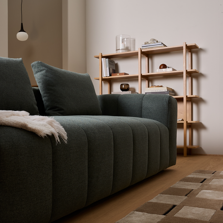 Mørkegrøn sofa i close-up med plaid, træreol med bøger og dekorationer i en skandinavisk stue.