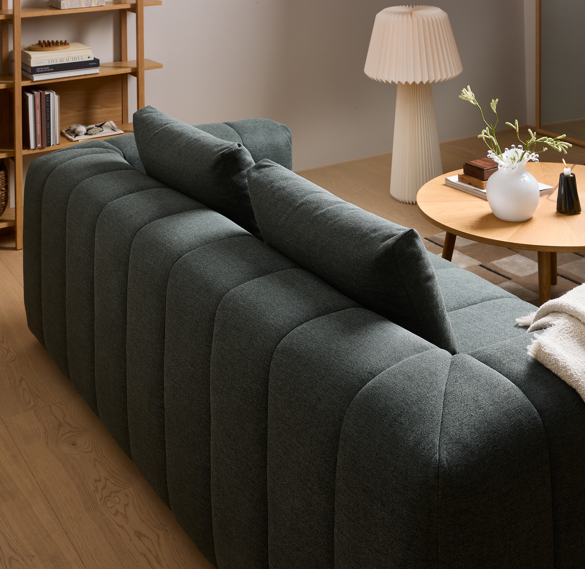 Mørkegrøn kanalquiltet sofa set bagfra i en lys stue med rundt træbord, hvid riflede vase med grene, et lys og en plisseret bordlampe.