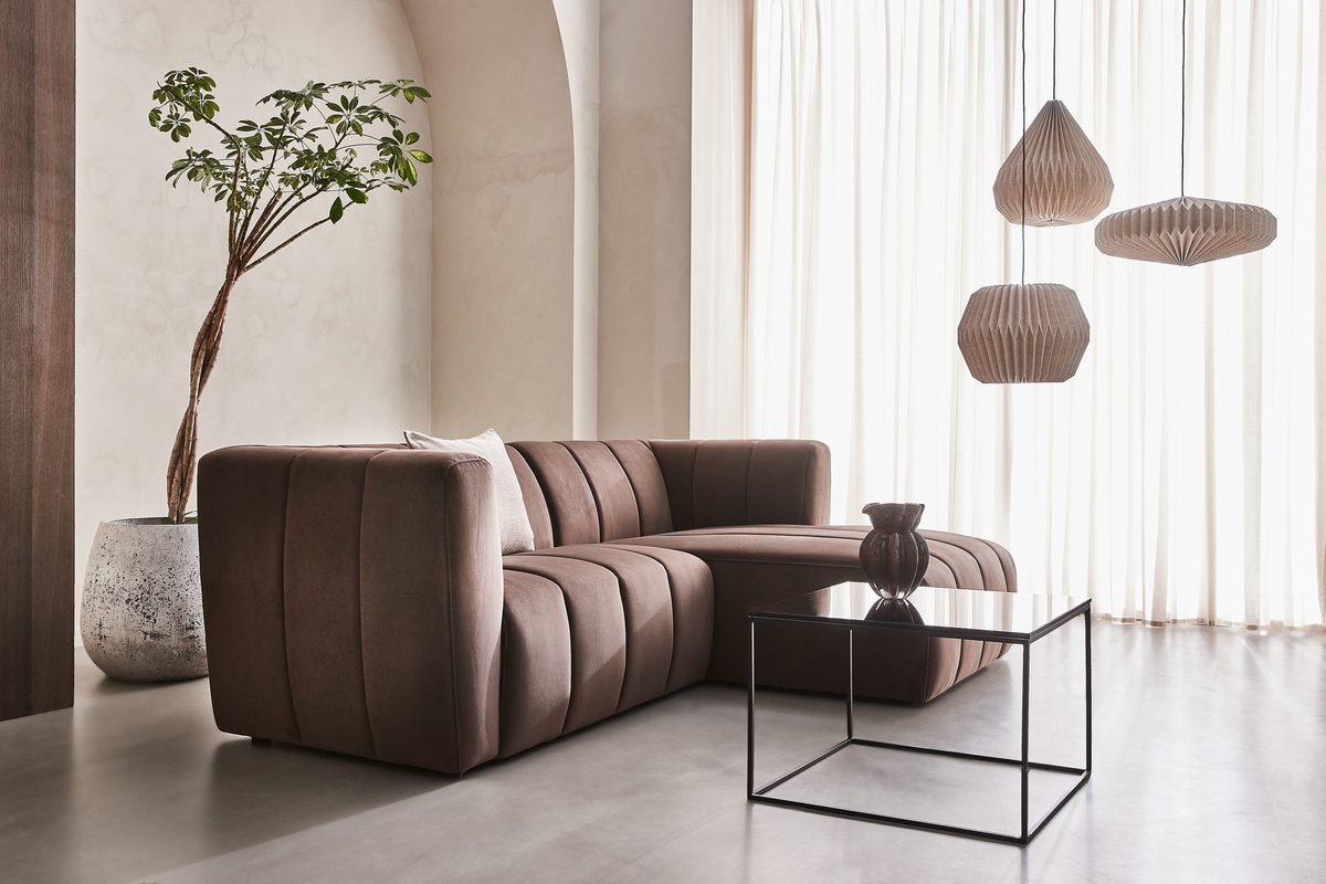 Minimalistisk stue med brun modulsofa, sort glas-sofabord, tre foldede pendellamper og en stor potteplante foran lyse gardiner.