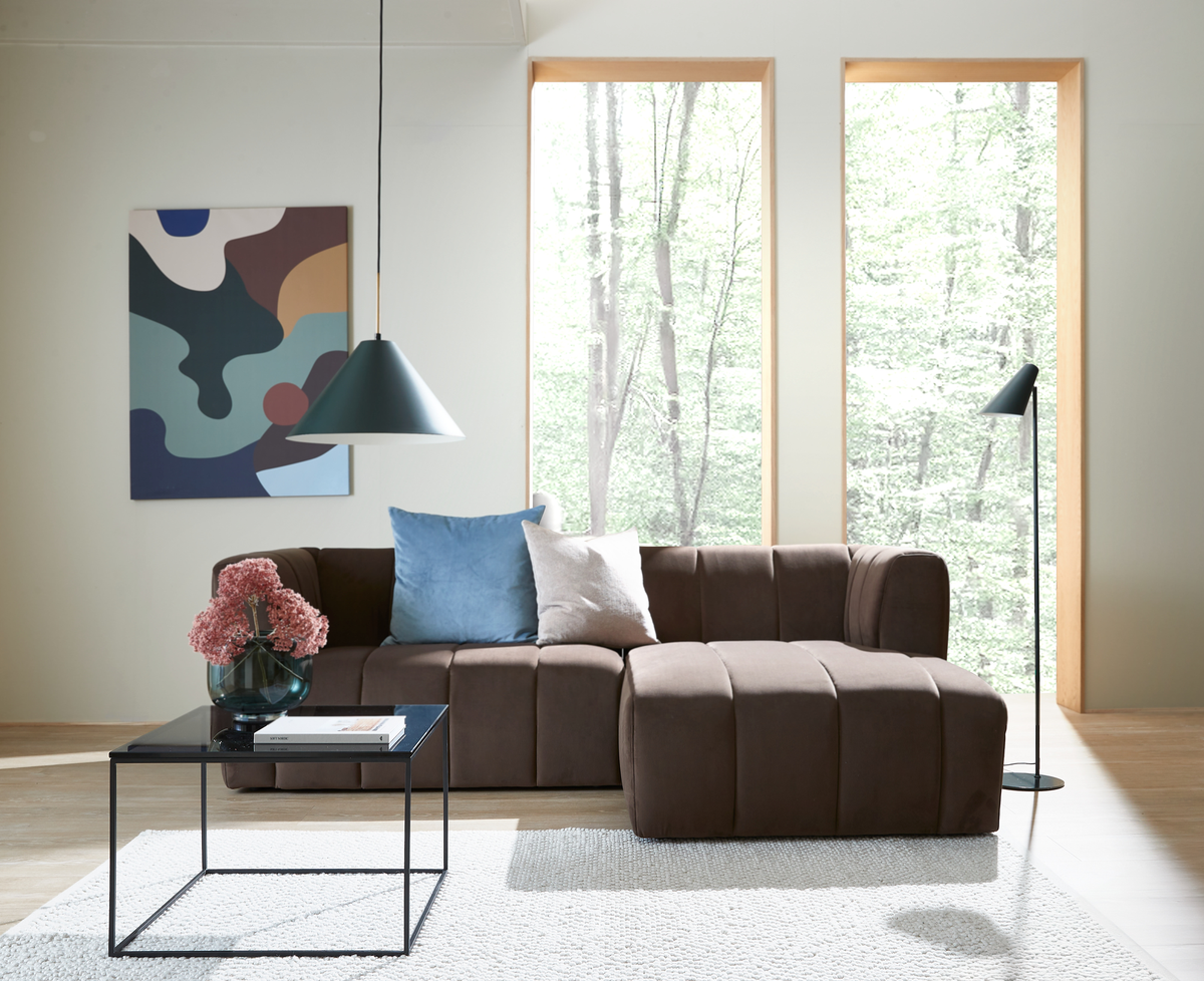 Lyst stue med brun modulsofa, blå og beige puder, sort sofabord med vase med lyserøde blomster, pendel- og gulvlampe samt store vinduer mod skov.