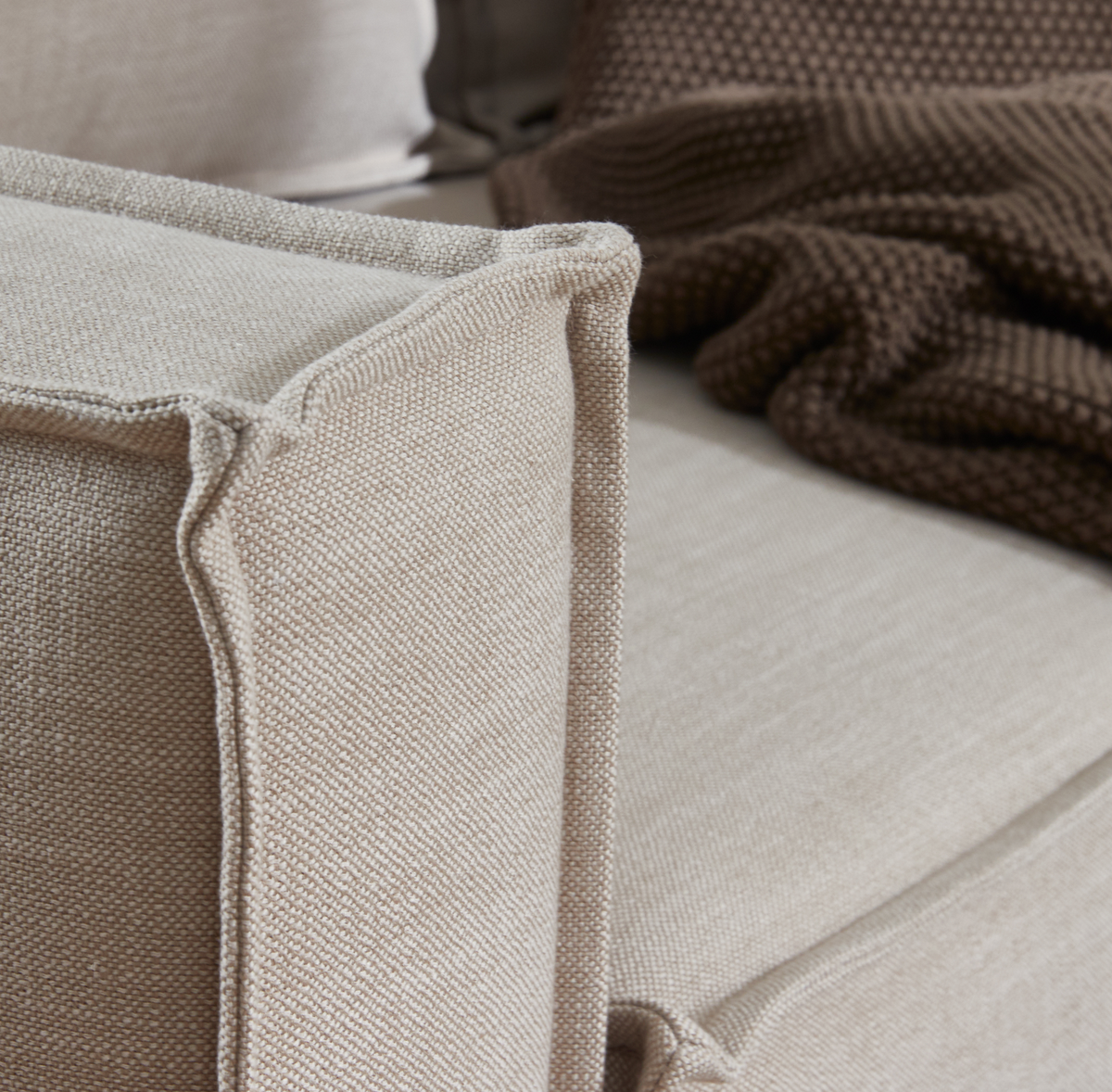 Nærbillede af beige stofsofa med markeret kantsyning og en brun strikket plaid i baggrunden.