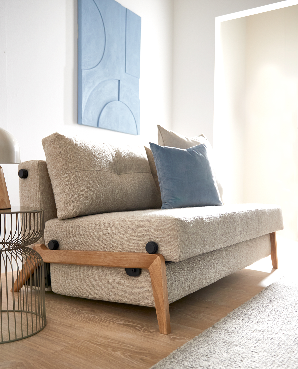 Beige sofa med træben og blå pude i en lys stue; abstrakt blå vægpanel og rundt metalbord ved siden af.