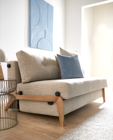 Beige sofa med træben og blå pude i en lys stue; abstrakt blå vægpanel og rundt metalbord ved siden af.