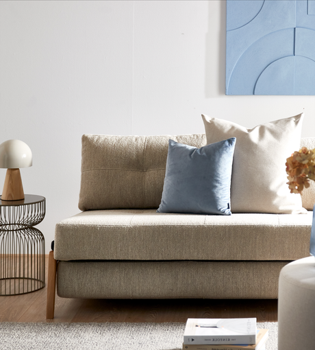 Beige sofa med blå og hvid pude, rund bordlampe og blå vægkunst; på tæppet ligger bøger, hvoraf en har titlen “The Kinfolk Home”.