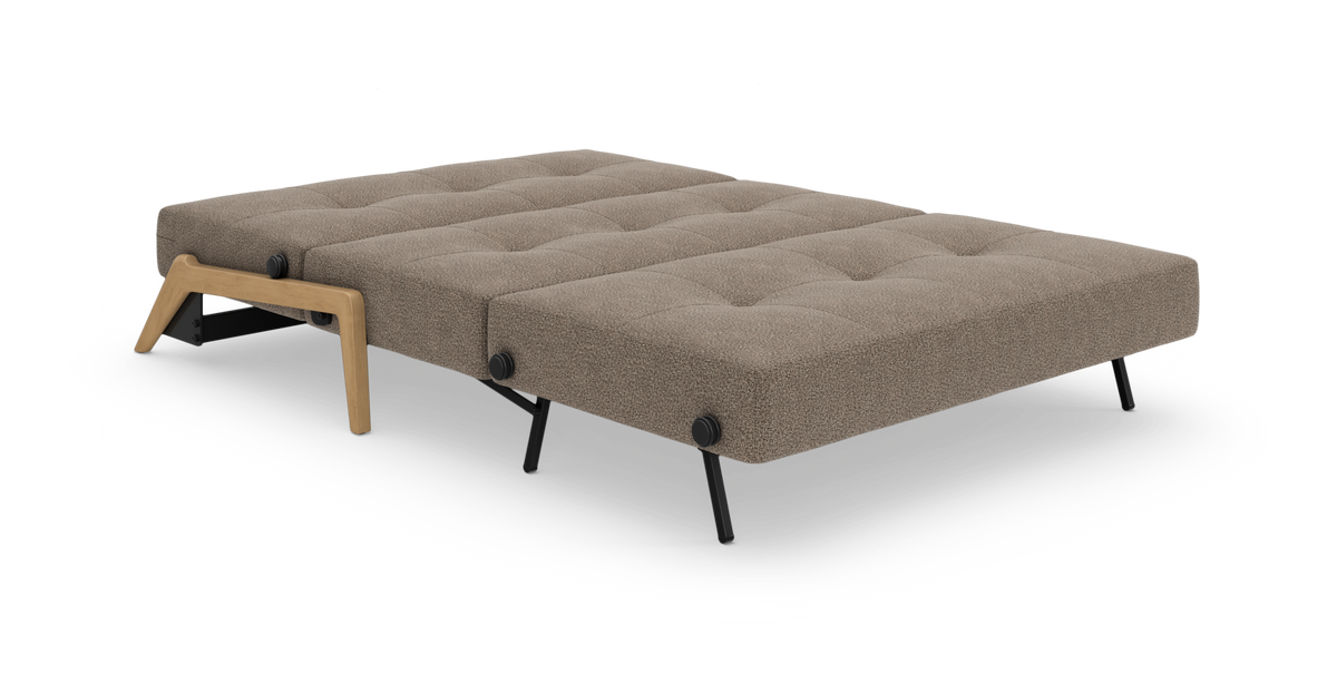 Sovesofa i udslået sengelayout, beige/taupe betræk med knapptuftet overflade, sorte metalben og træben i egetræslook, frilagt på hvid baggrund.