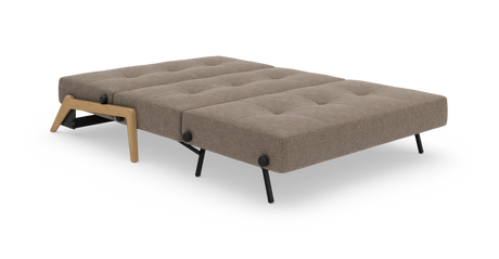 Sovesofa i udslået sengelayout, beige/taupe betræk med knapptuftet overflade, sorte metalben og træben i egetræslook, frilagt på hvid baggrund.