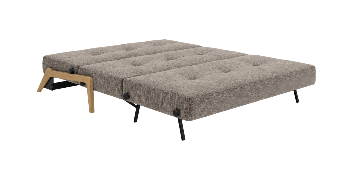 Gråpolstret sovesofa i udfoldet position med tuftede puder, sorte metalben og trædetalje ved armlæn, fritlagt mod hvid baggrund i skrå frontvinkel.