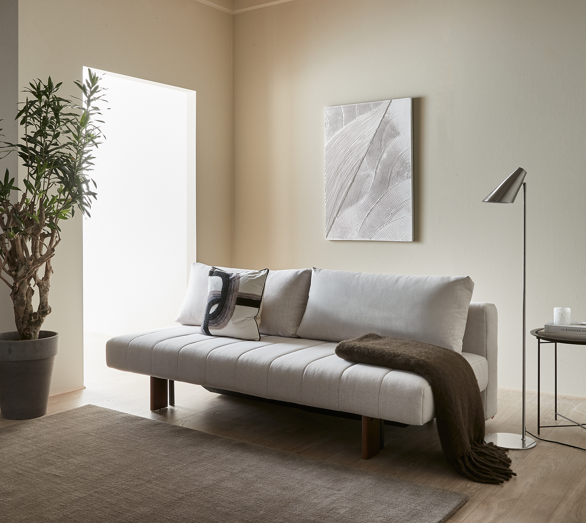 Lyst minimalistisk stue med beige sofa med puder og brun plaid, gulvlampe og sidebord med lys og bogen Kinfolk, vægkunst og stor plante ved lyst vindue.