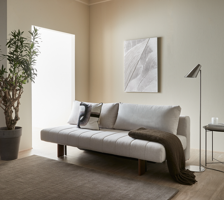 Lyst minimalistisk stue med beige sofa med puder og brun plaid, gulvlampe og sidebord med lys og bogen Kinfolk, vægkunst og stor plante ved lyst vindue.