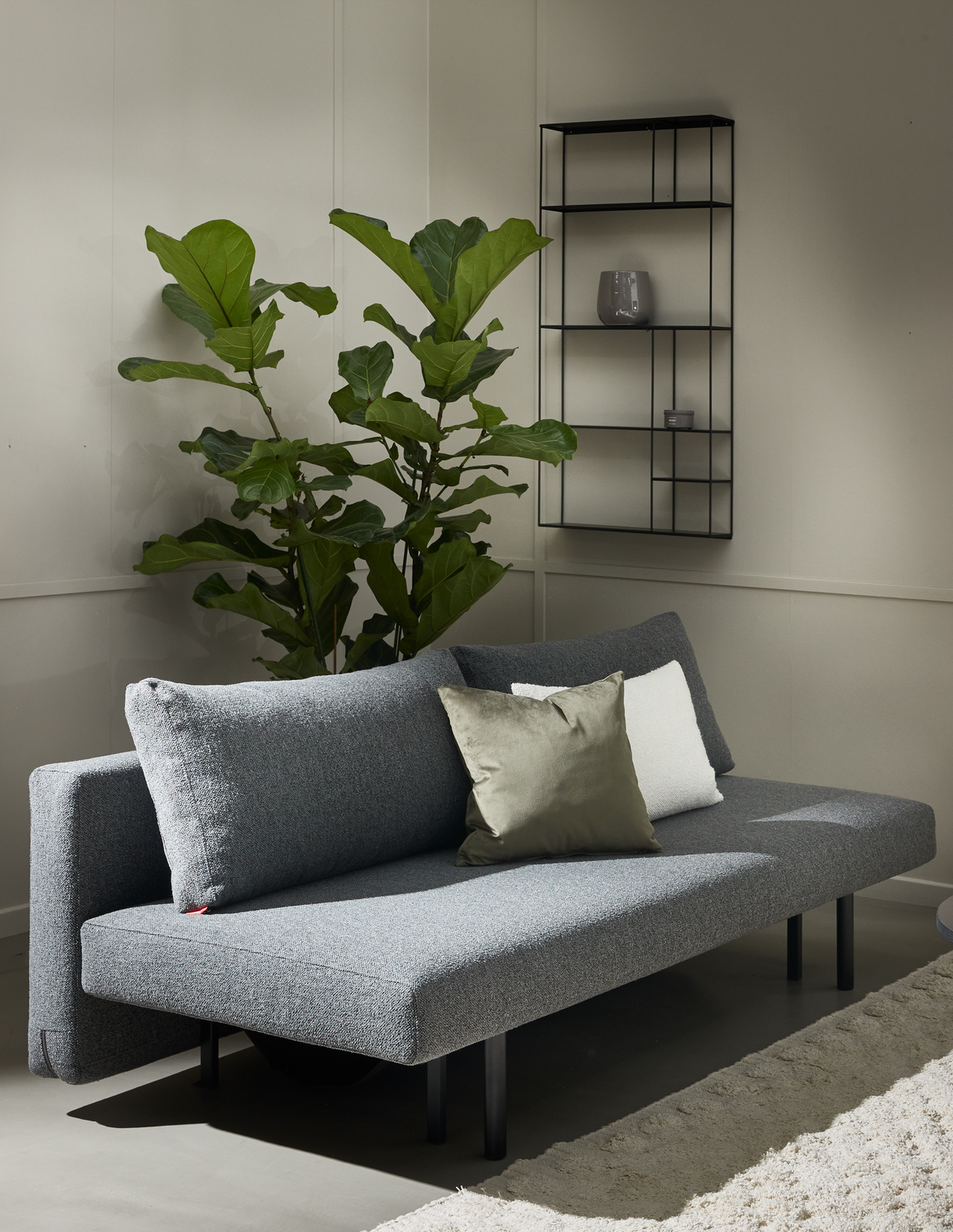 Grå minimalistisk sofa med grøn og hvid pude i en lys stue, stor violinfigen og sort væghylde over et lyst tæppe.