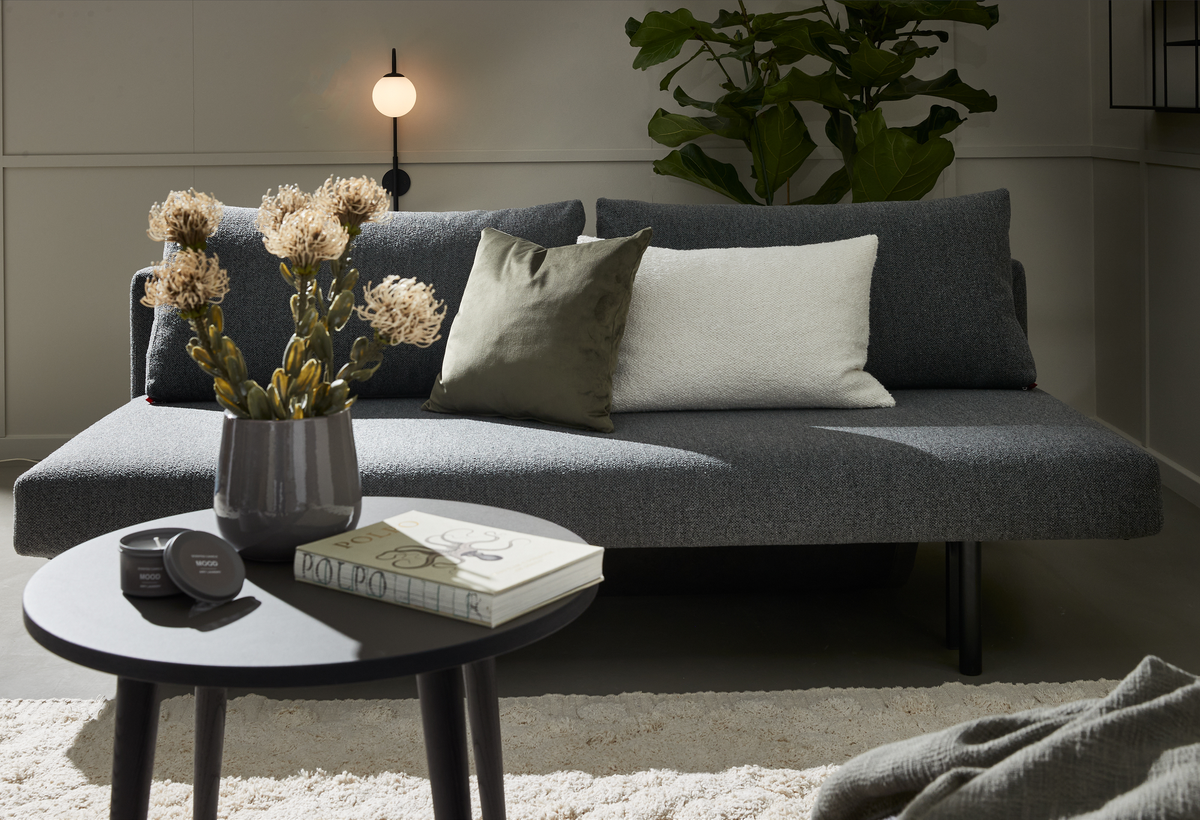 Grå sofa med hvide og grønne puder i en stue; rundt sort sofabord med vase med tørrede blomster, to små runde dåser med “MOOD” og en bog med “POPO”; tændt væglampe, stor grøn plante og lys måtte.