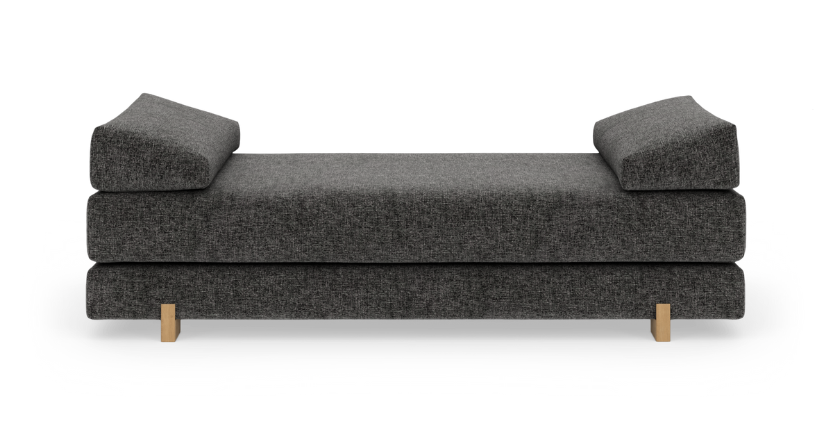 Mørkegrå daybed/sofa med to skrånende pudestøtter og lyse træben, set forfra på hvid baggrund.