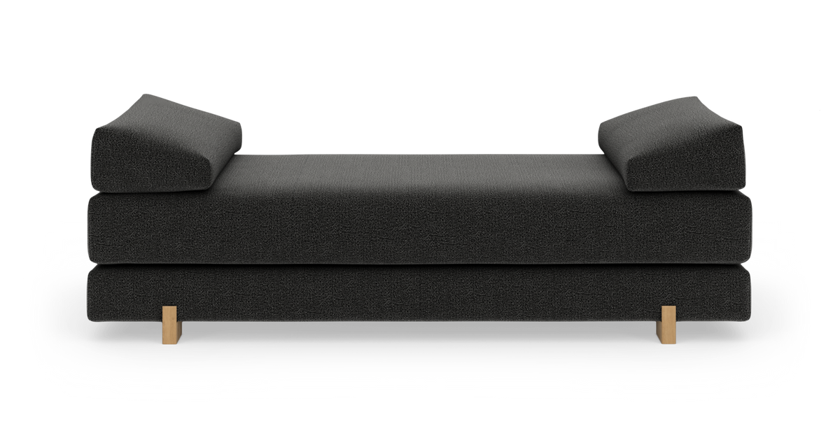 Mørkegrå daybed/sofa med to kileformede armlæn og lyse træben, frilagt på hvid baggrund i frontvisning.