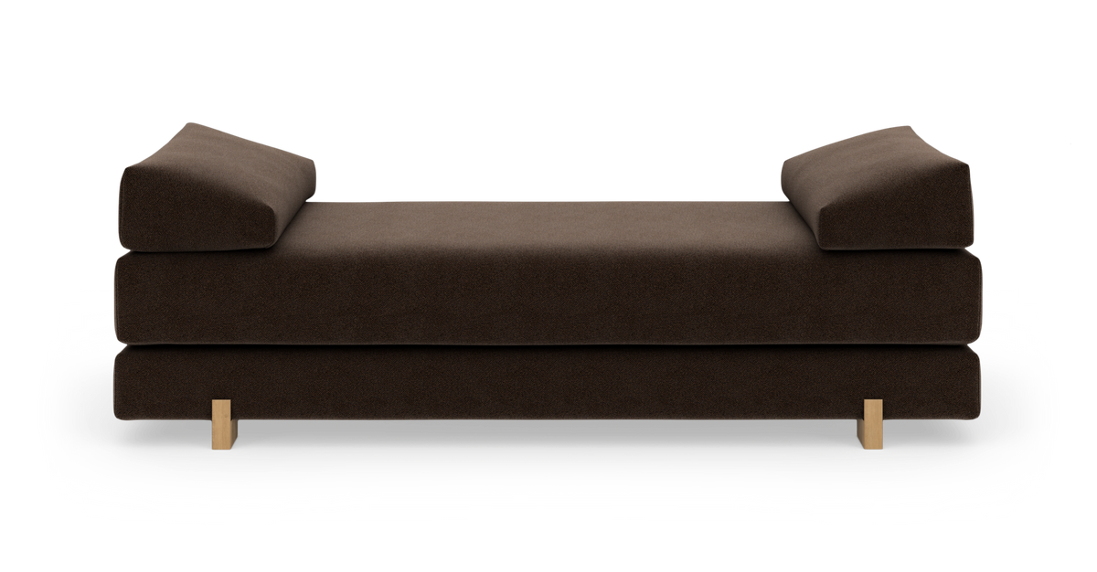 Brun daybed med skrå armlæn og lyse træben, fotograferet forfra på hvid baggrund.