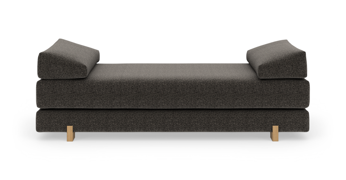 Mørkegrå daybed med to vinklede bolsterpuder og lyse træben, frilagt på hvid baggrund i frontvisning.