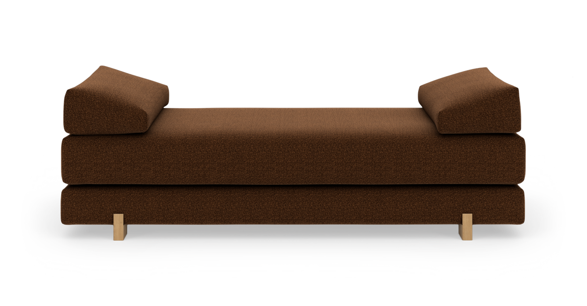 Brun daybed/sofa med vinklede puder i begge ender og lyse træben, fotograferet lige forfra på hvid studiebaggrund.