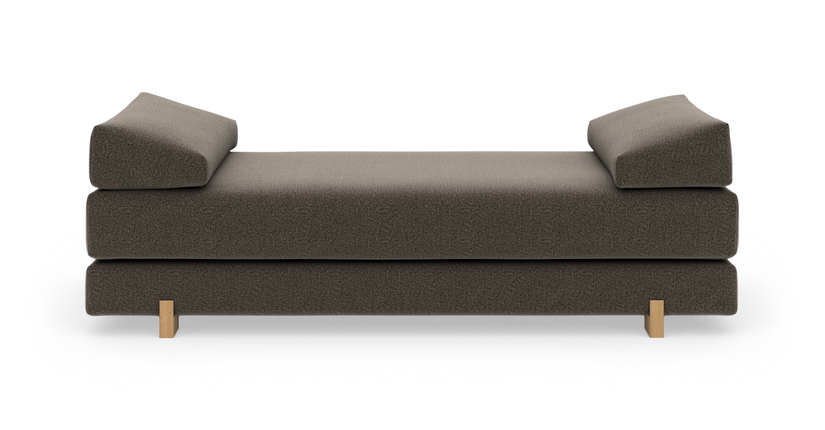 Moderne daybed i brunmeleret tekstil med to kileformede armlæn og lyse træben, set forfra i studiemiljø.