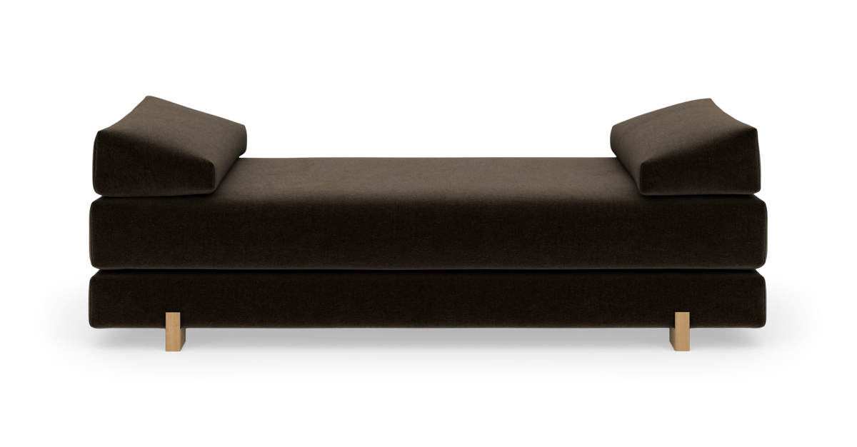 Mørkebrun polstret daybed med vinklede armpuder i begge ender og lyse træben, frontvisning mod hvid studiebaggrund.