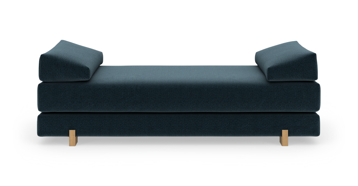 Mørk blågrøn daybed med to kileformede endepuder og lyse træben, frilagt på hvid baggrund set forfra.