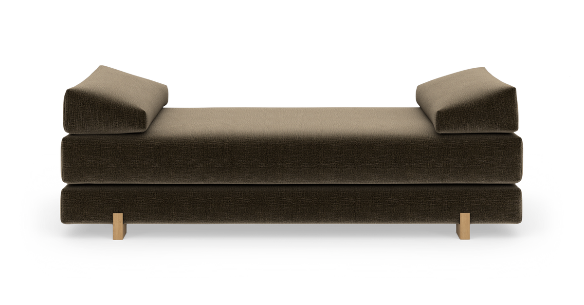 Moderne daybed/sofa i mørkebrun tekstil med to kileformede armlænspuder og lyse træben, frilagt på hvid baggrund.