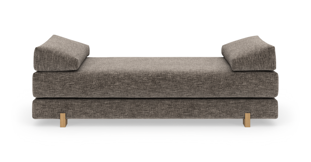 Grå polstret daybed med to skrå puder og lyse træben, fotograferet lige forfra på hvid baggrund.