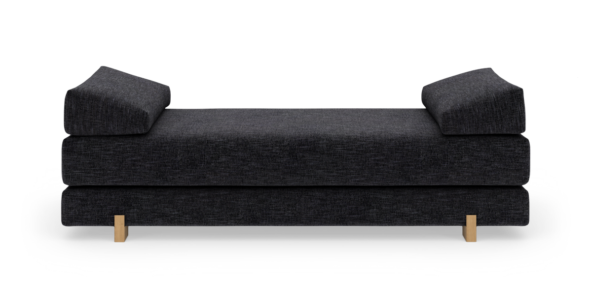 Mørkegrå daybed/sofa med to kileformede armlænspuder og lyse træben, fritlagt på hvid baggrund.