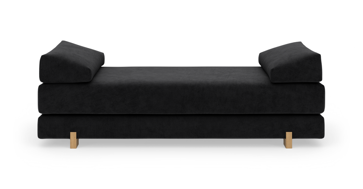 Sort daybed med vinklede armpuder og lyse træben, fotograferet forfra.
