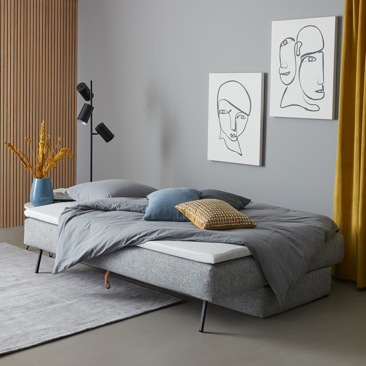 Moderne soveværelse med grå polstret seng og gråt sengetøj, blå og gul pyntepude, sort gulvlampe, vægkunst med linjetegninger, gule gardiner og en vase med tørrede planter på en grå måtte.