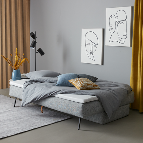 Moderne soveværelse med grå polstret seng og gråt sengetøj, blå og gul pyntepude, sort gulvlampe, vægkunst med linjetegninger, gule gardiner og en vase med tørrede planter på en grå måtte.