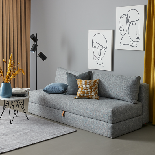 Moderne grå sofa med puder, lille rundt sidebord med blå vase og tørrede grene, sort gulvlampe, to linjetegninger på grå væg og gule gardiner i en skandinavisk stue.