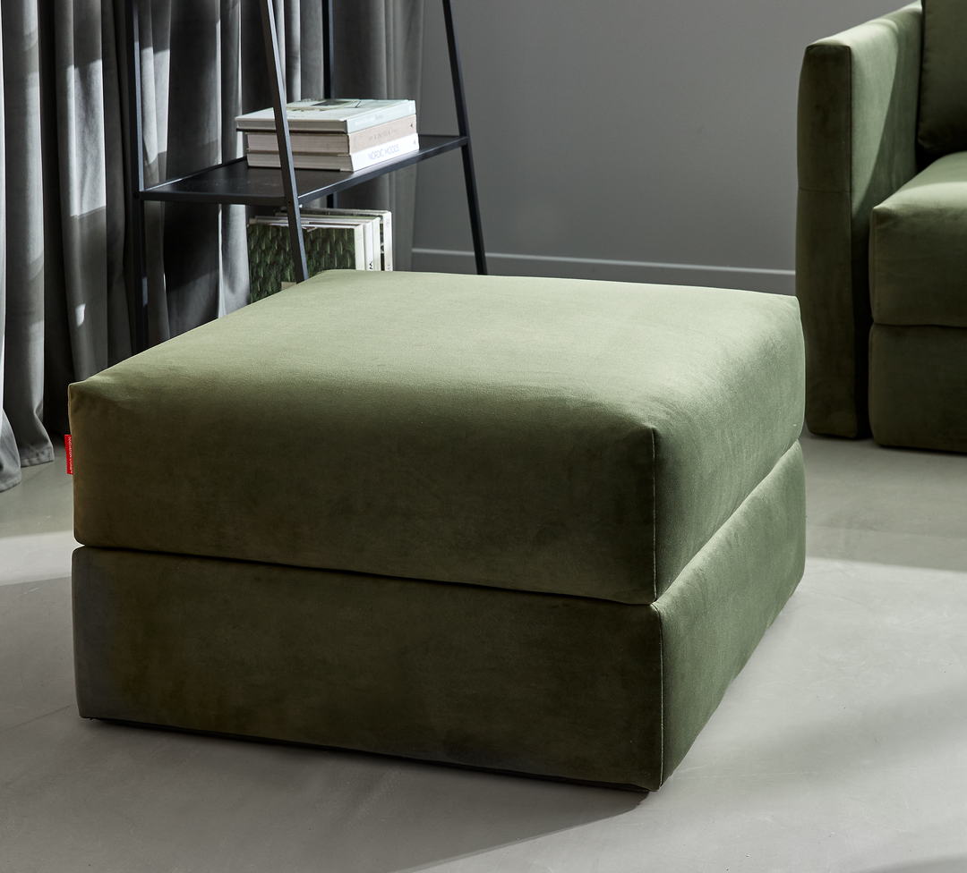 Olivengrøn polstret ottoman i moderne stue ved siden af sofa og reol med bøger i dagslys.