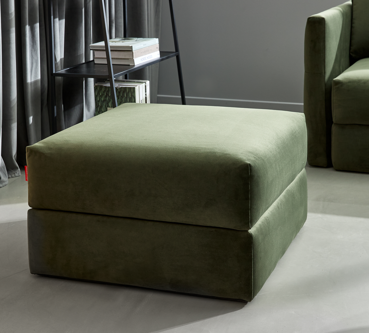 Olivengrøn polstret ottoman i moderne stue ved siden af sofa og reol med bøger i dagslys.