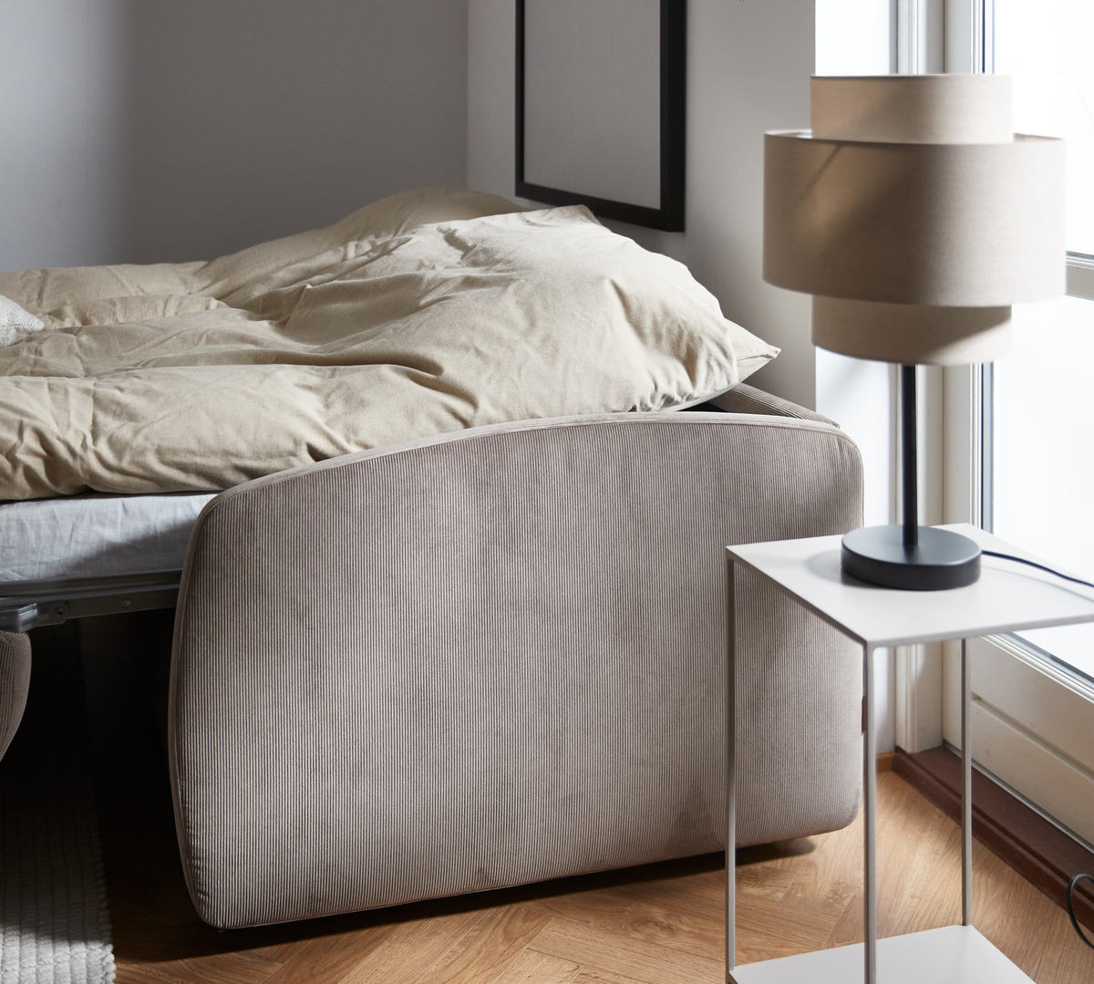 Lyst soveværelse med beige sengegavl, redt seng og moderne bordlampe på hvidt sidebord ved vindue.