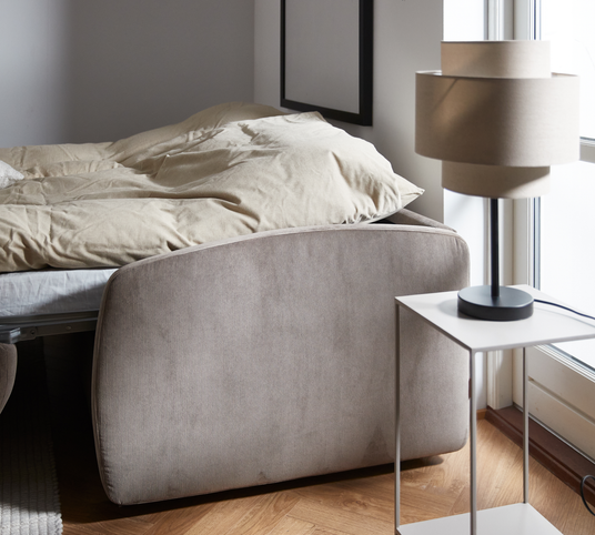 Lyst soveværelse med beige sengegavl, redt seng og moderne bordlampe på hvidt sidebord ved vindue.